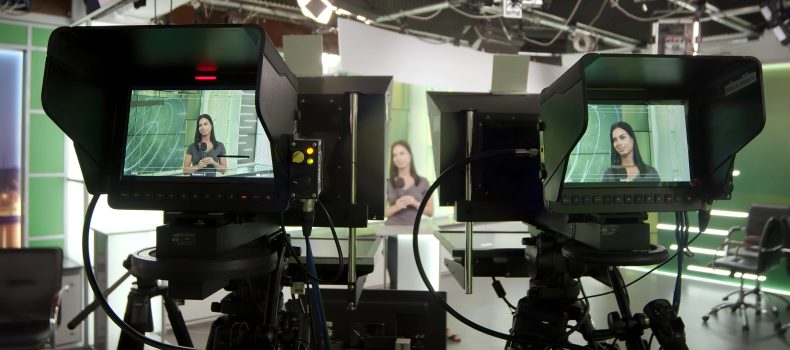 Two broadcast television cameras on tripods filming a presenter in a studio