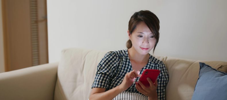 Woman sitting on a sofa at home in the evening watching a video podcast on her phone