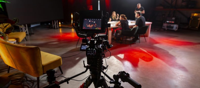 A television studio camera filming an interview set where a host and guests sit at a lit table