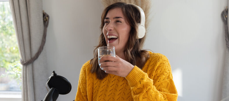 Podcaster with headphones laughing and sipping water at her microphone before recording